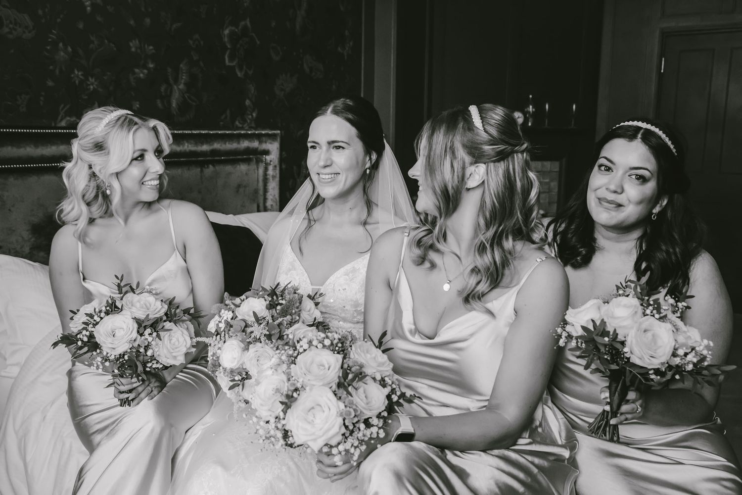 Black and white photo of a bride in veil with three bridesmaids in satin dresses holding floral bouquets.