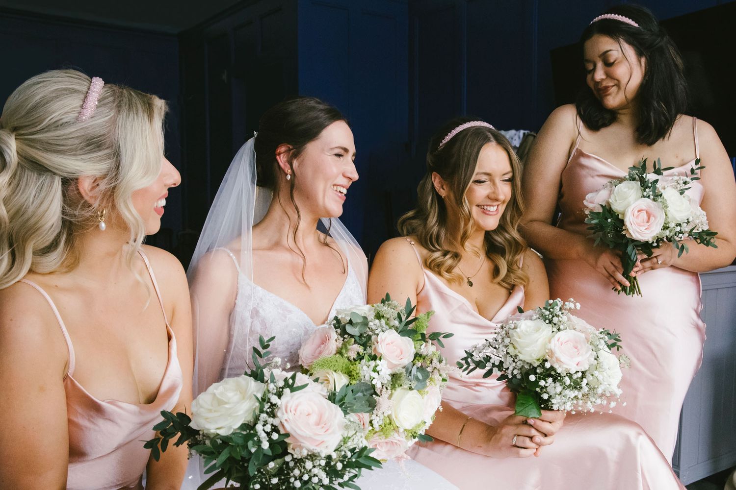 Bride and bridesmaids in blush pink dresses laughing together holding white floral bouquets with greenery.