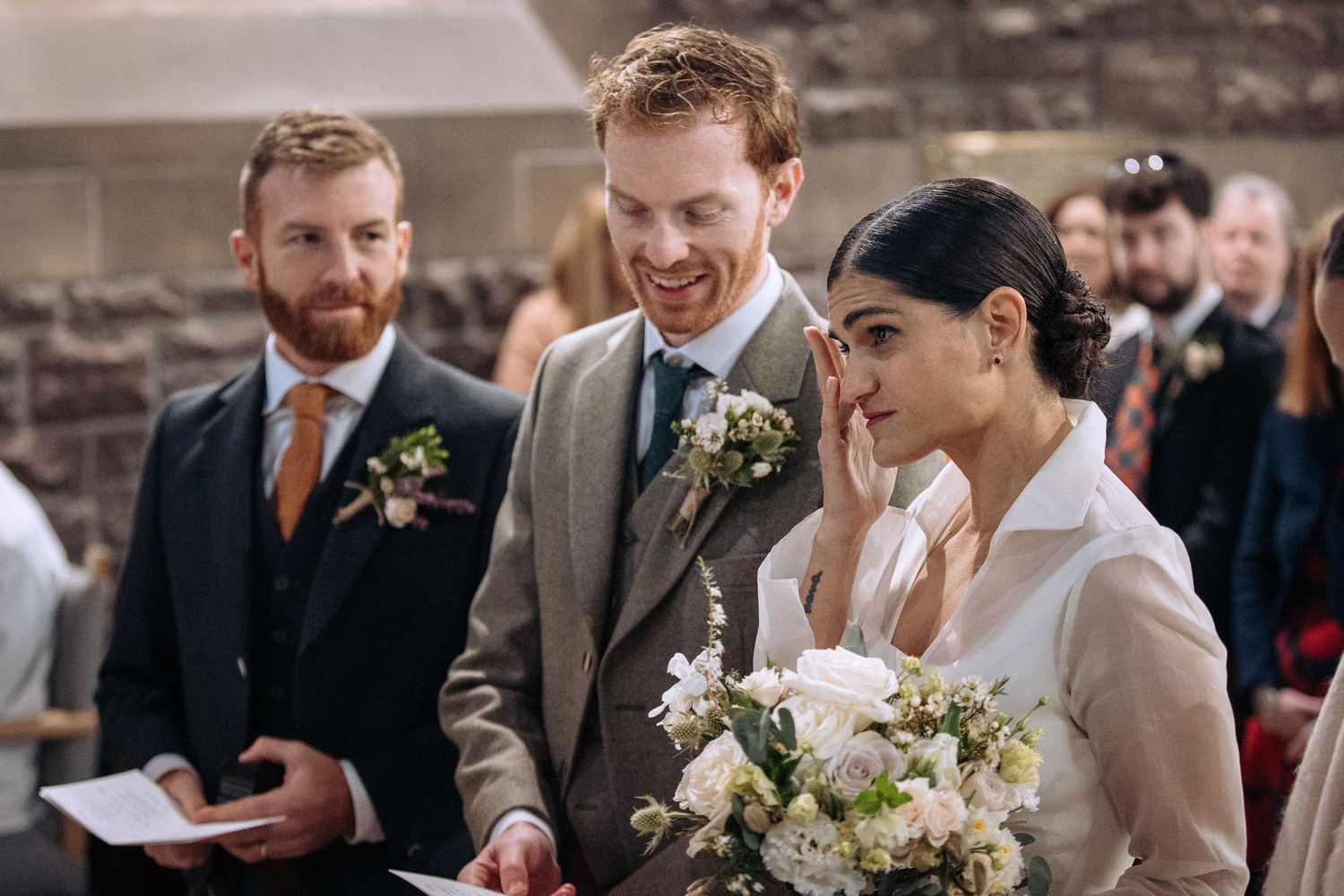 Emotional bride wiping tears holding bouquet beside smiling groom in grey suit during wedding ceremony.