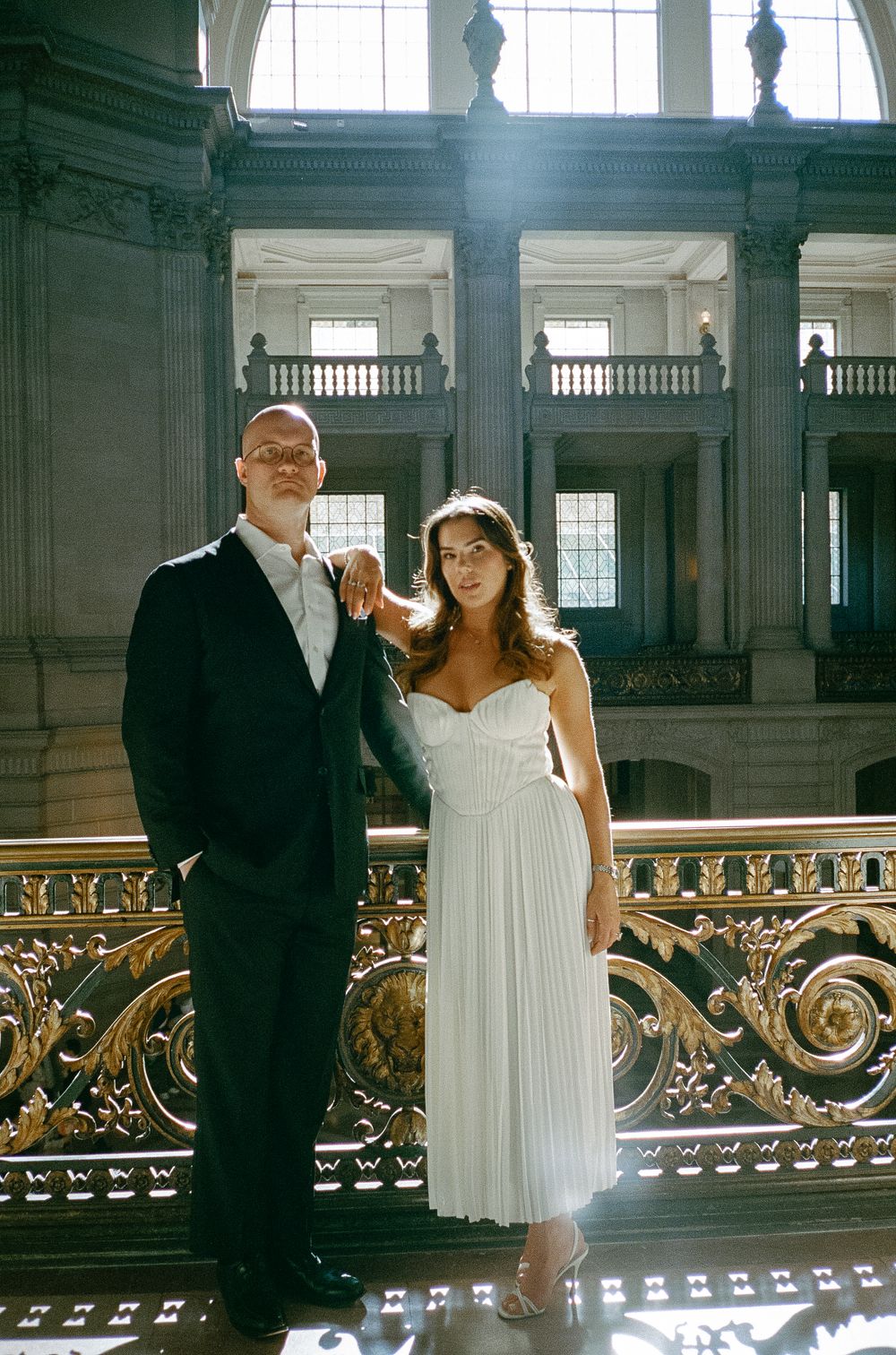 Couple in wedding attire posing on ornate golden staircase balcony inside San Francisco City Hall.