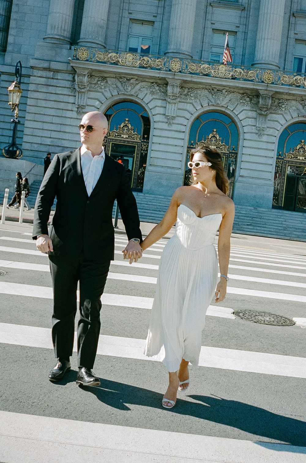 Couple holding hands crossing a crosswalk outside San Francisco City Hall on a sunny wedding day.