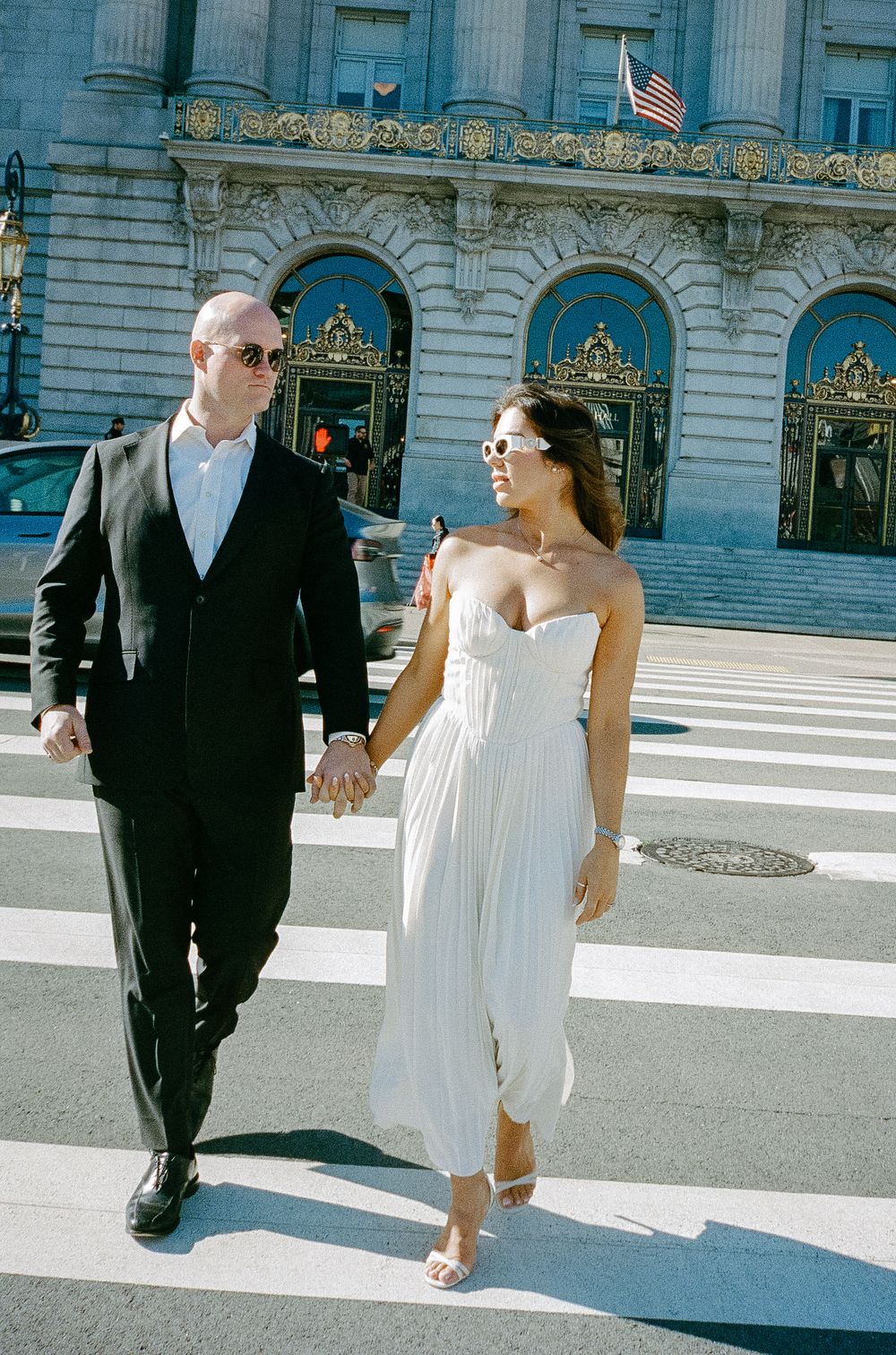 Newlyweds in sunglasses walking hand in hand across sunny crosswalk outside San Francisco City Hall.