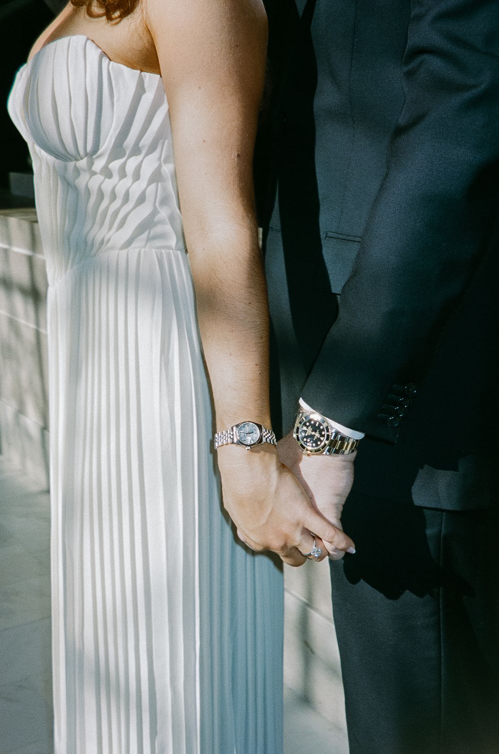 Close-up of wedding couple holding hands, bride in white pleated gown, both wearing silver watches.