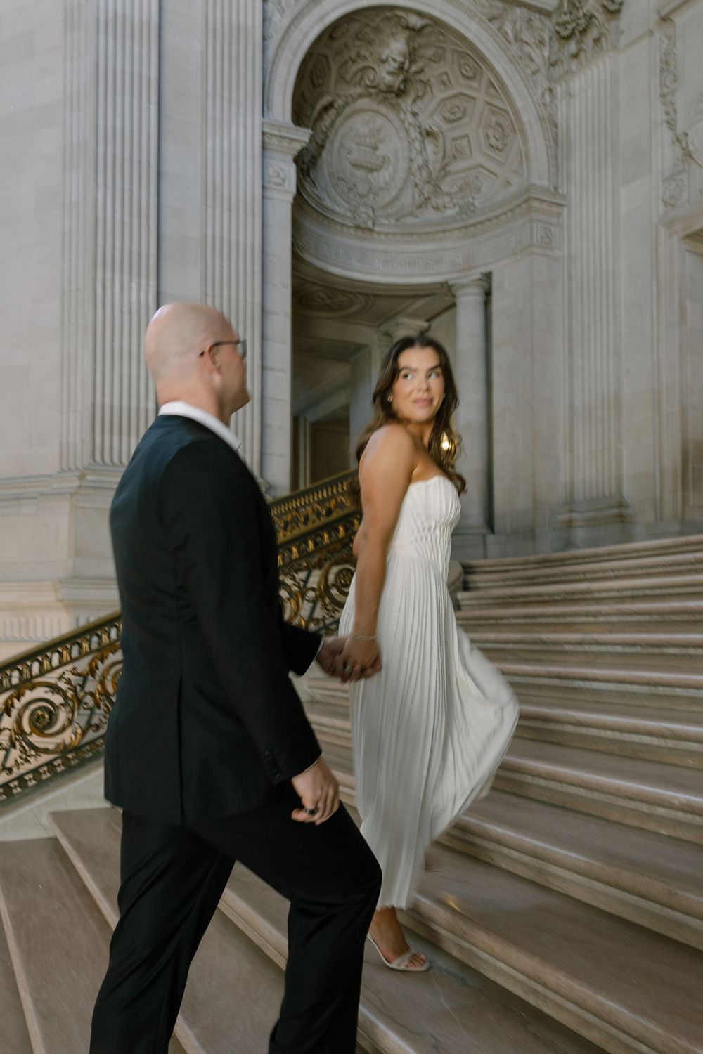 Couple in wedding attire holding hands on grand marble staircase, bride in white pleated gown looking back at camera.