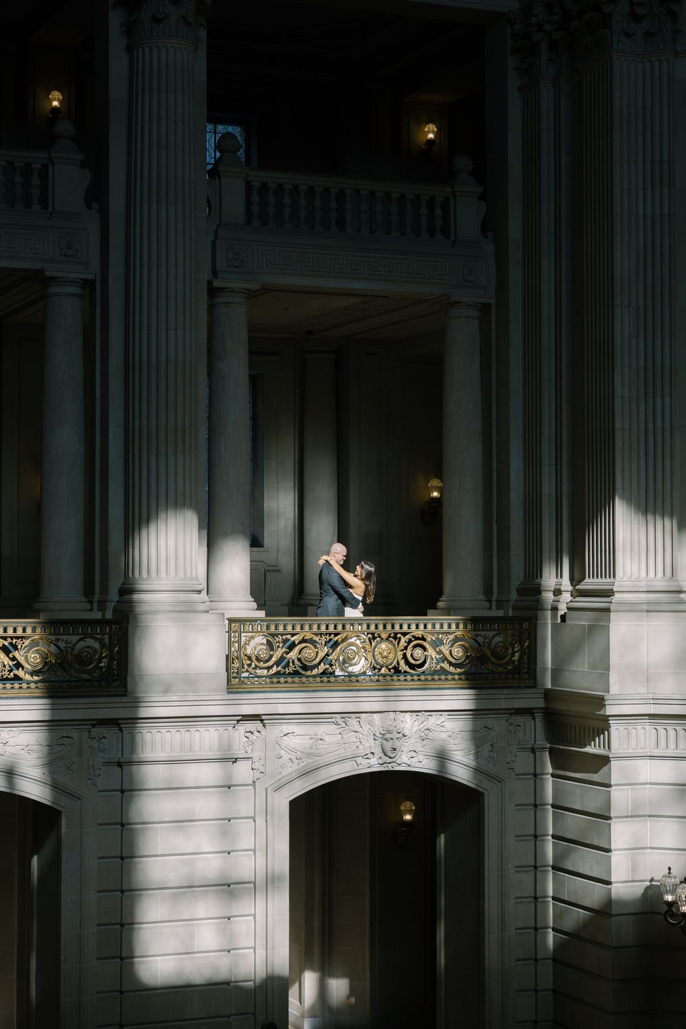 Couple embraces on decorative balcony between tall columns of elegant neoclassical building interior.