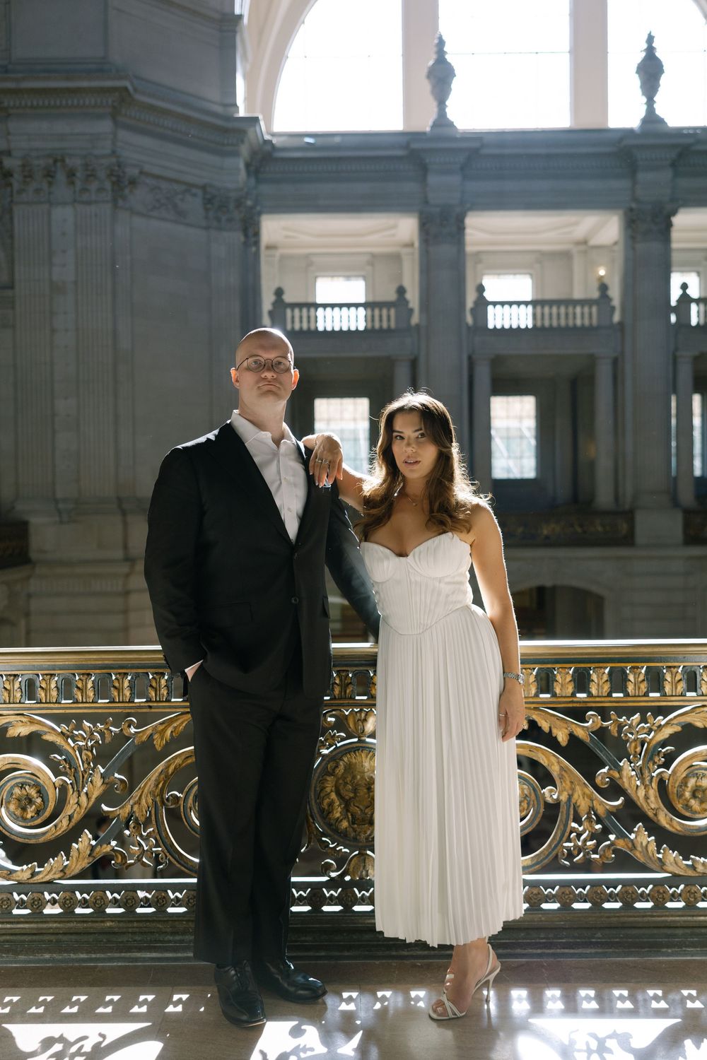 Bride and groom posing by ornate gold railing in grand wedding venue interior.