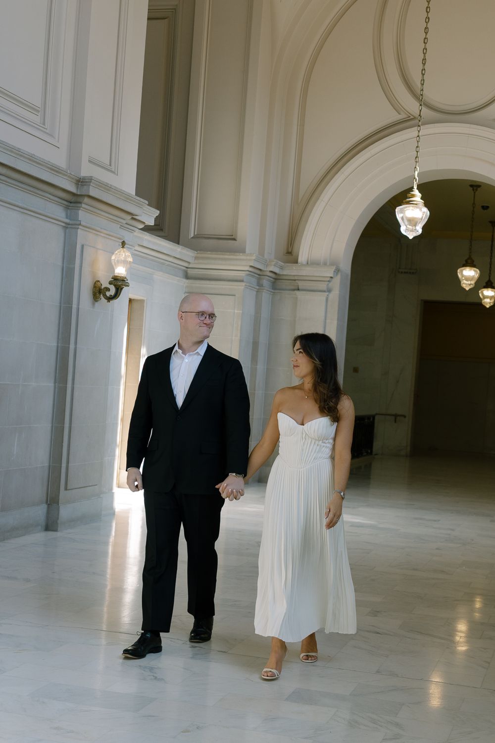 Newlyweds walking hand in hand through elegant marble hallway of San Francisco City Hall in wedding attire.