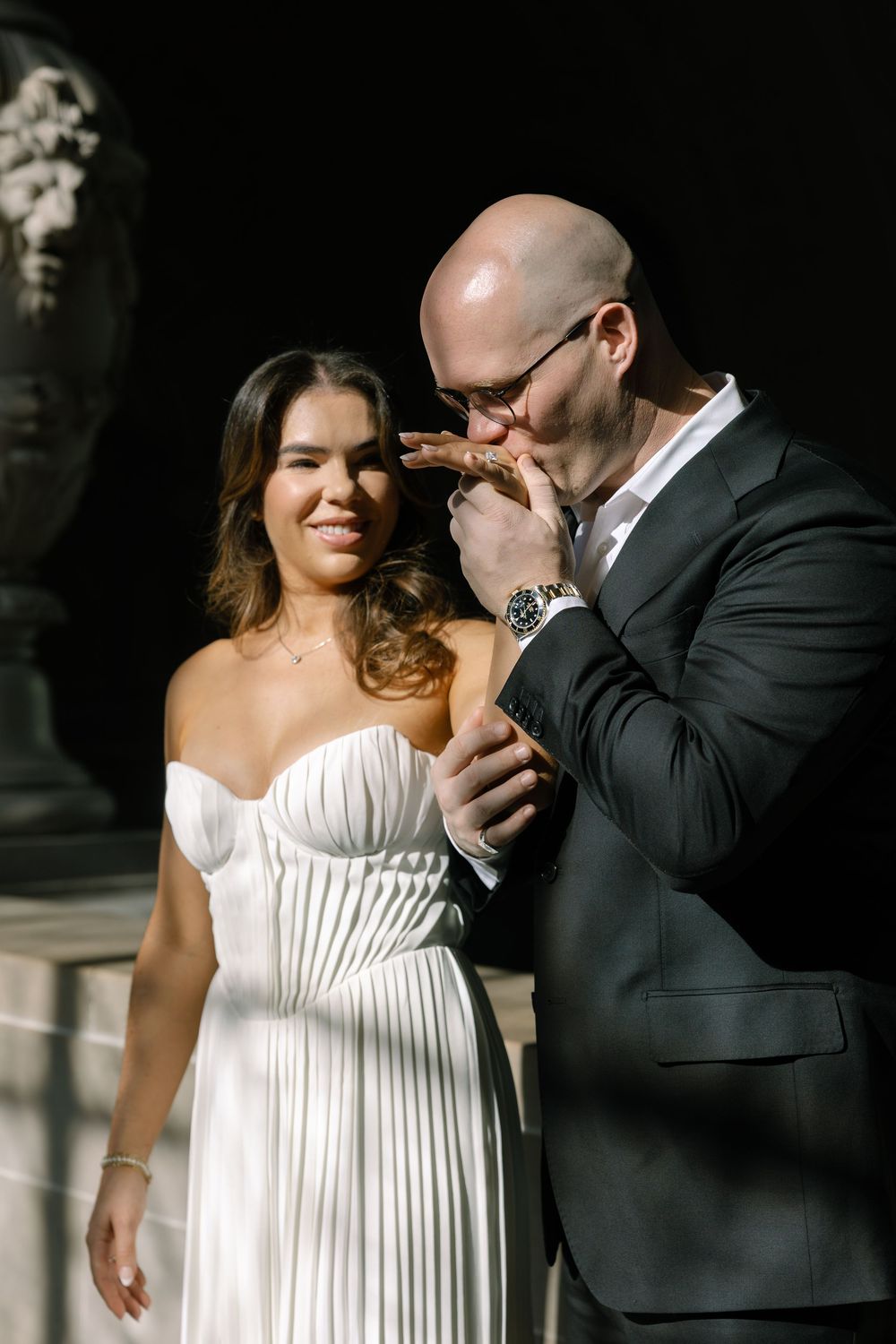 Groom in black suit kisses bride's hand as she smiles in pleated white wedding gown outdoors.
