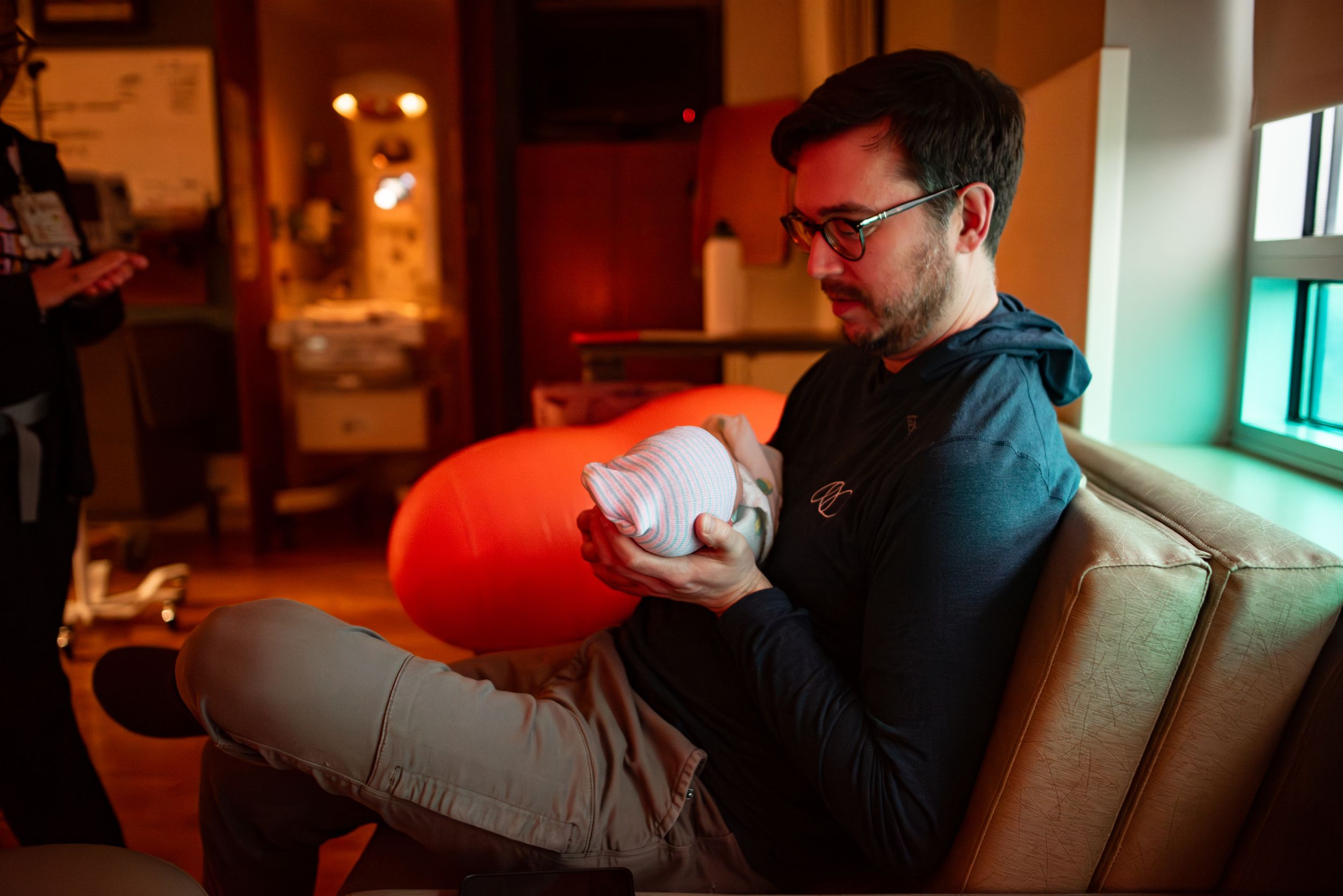 New father sitting on couch holding swaddled newborn baby in dimly lit birth suite.