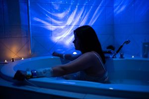 Woman relaxing in a dimly lit bathtub with blue ambient lighting and candles during a water birth preparation.