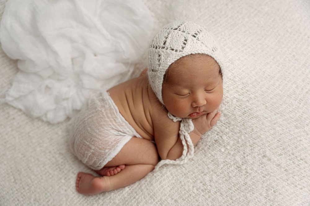 Sleeping newborn baby wearing a white knit bonnet, wrapped in cream fabric on a soft white blanket.