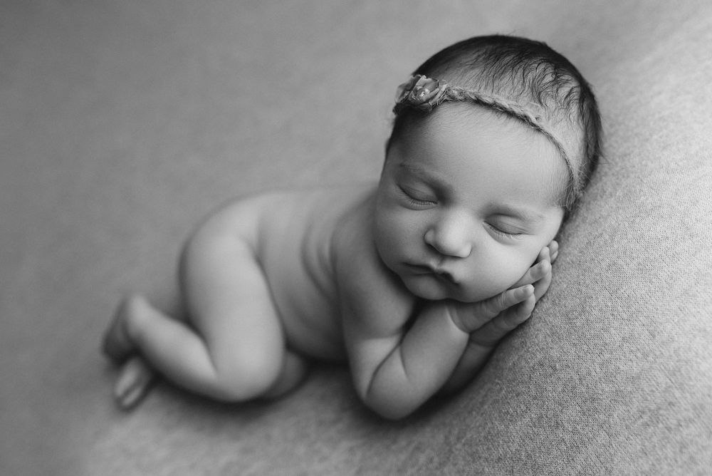 Black and white newborn photo of a sleeping baby girl with floral headband, curled up peacefully on a soft blanket.