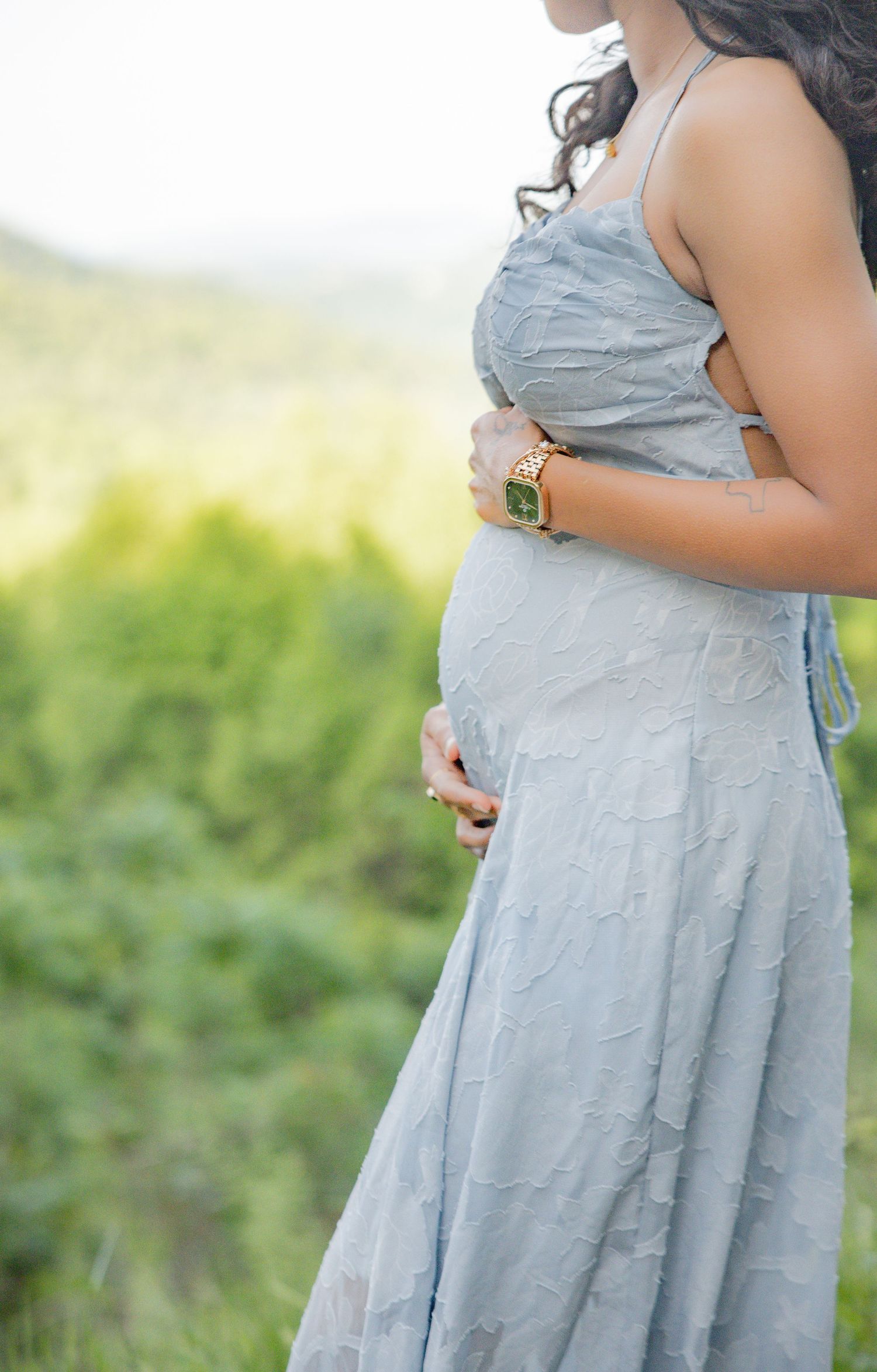 Pregnant woman in light blue dress holding baby bump outdoors with green hills in background.
