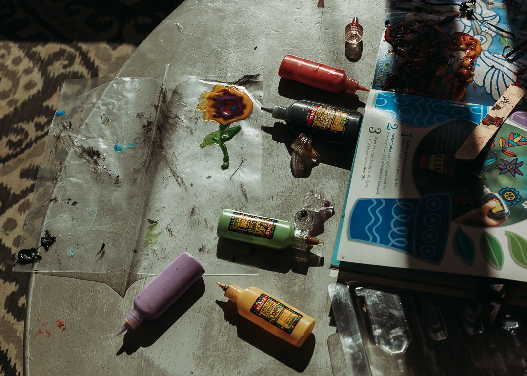 Assorted paint bottles and art supplies scattered on a glass table in a sunlit creative workspace.