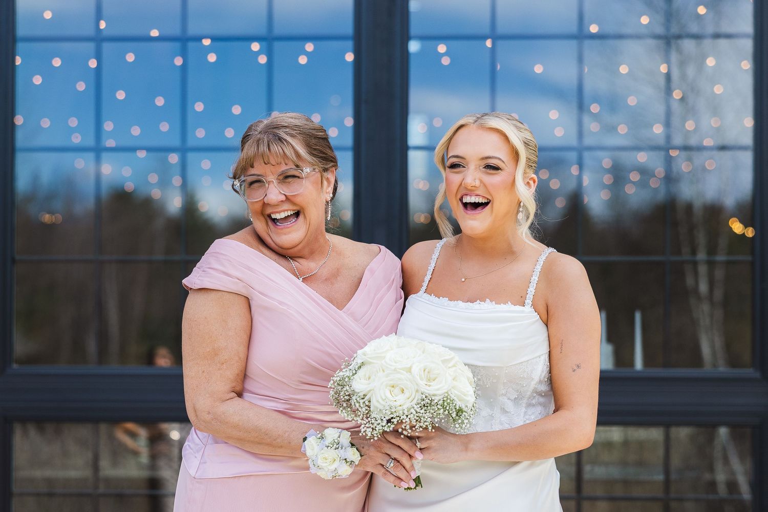 Bride in white gown holding white bouquet laughs with older woman in pink dress before large window.