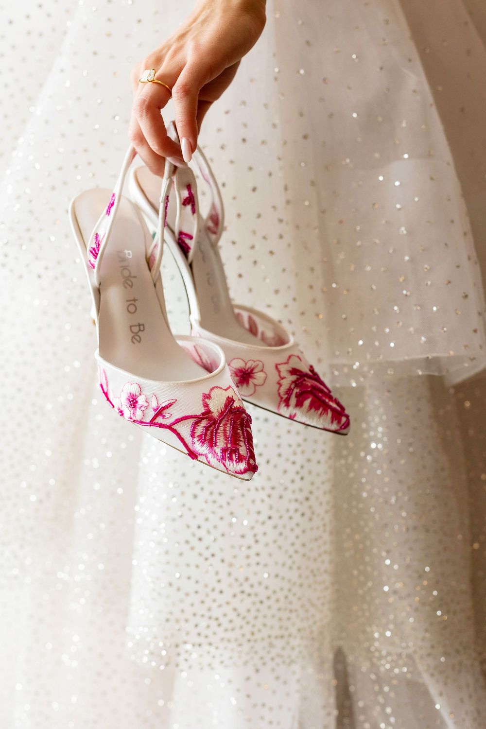 Bride holding white slingback heels with pink floral embroidery beside sparkly wedding gown.