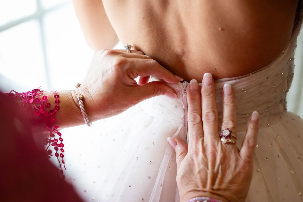 Hands fastening buttons on the back of a white wedding gown with delicate pearl details.