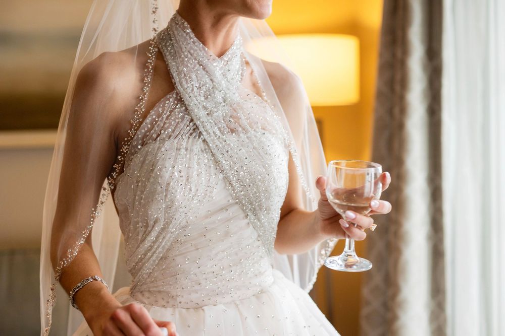Bride in beaded halter wedding gown holding a crystal glass of champagne by a window.