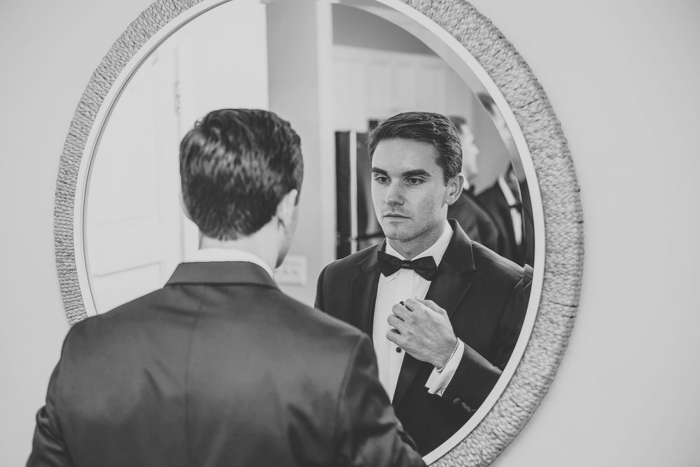 Groom in tuxedo adjusting bow tie while looking at reflection in ornate round mirror.