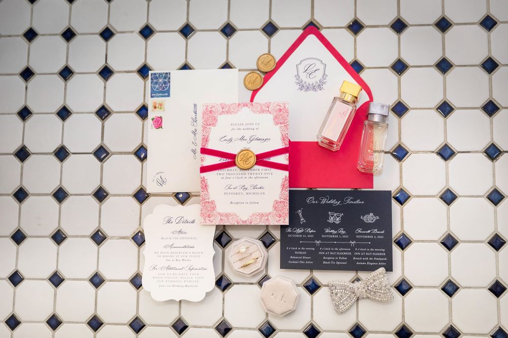 Elegant wedding invitation suite with red accents, gold wax seal, and perfume bottle on black and white tile.