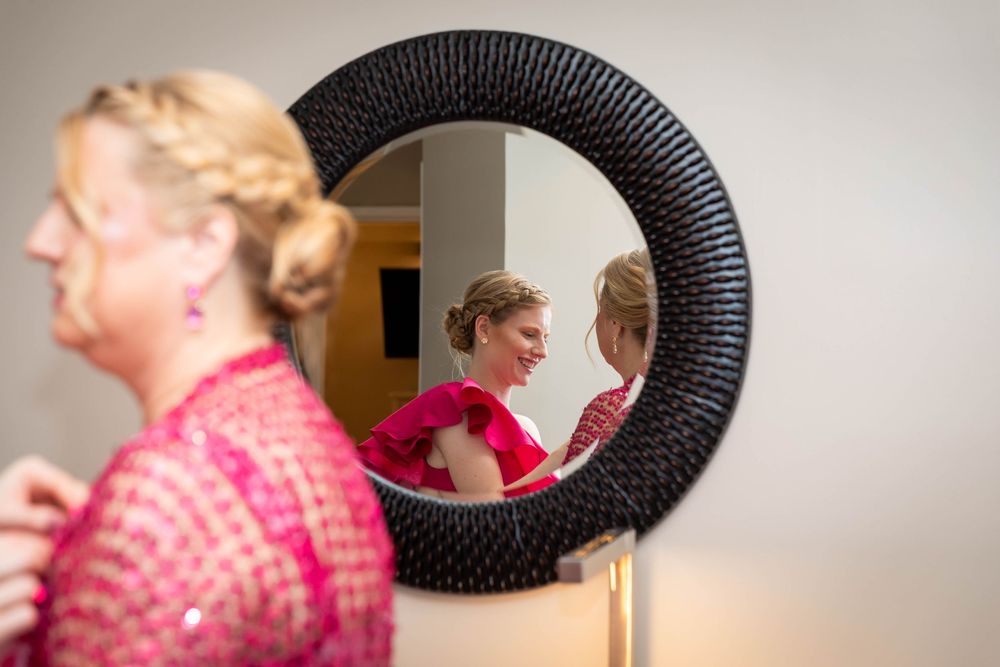 Bride smiling at her reflection in a round black mirror while woman in pink dress stands nearby.