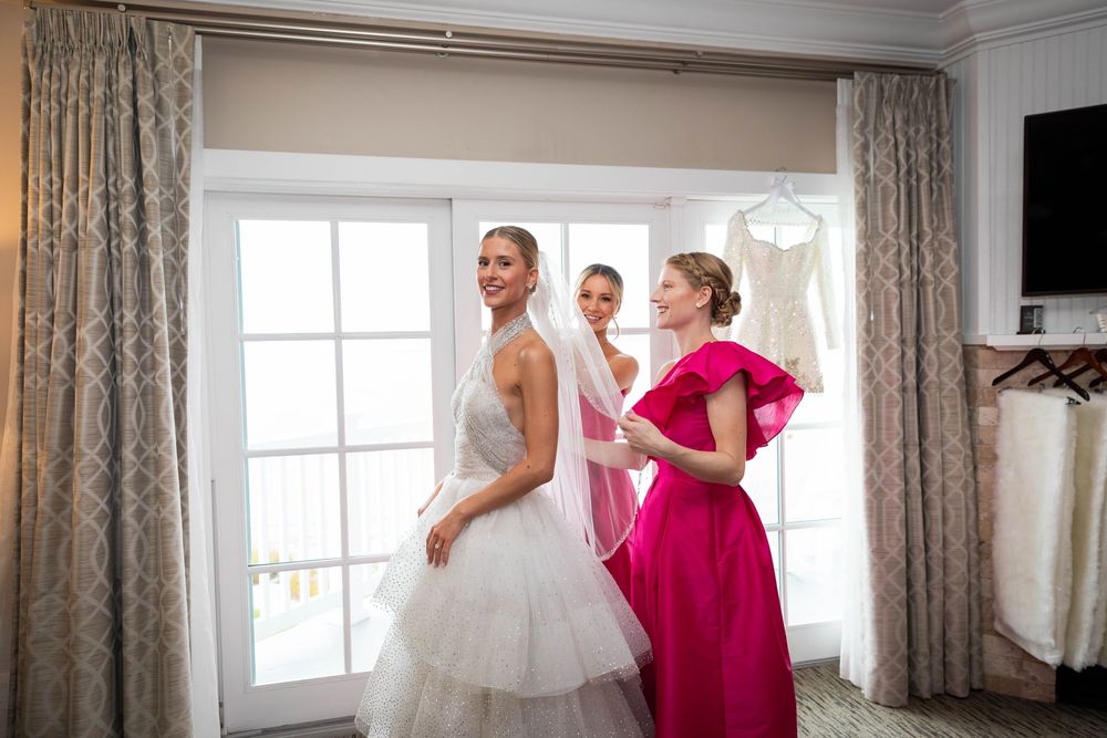 Bride in layered white gown and veil gets ready with bridesmaids in fuchsia dresses by window.