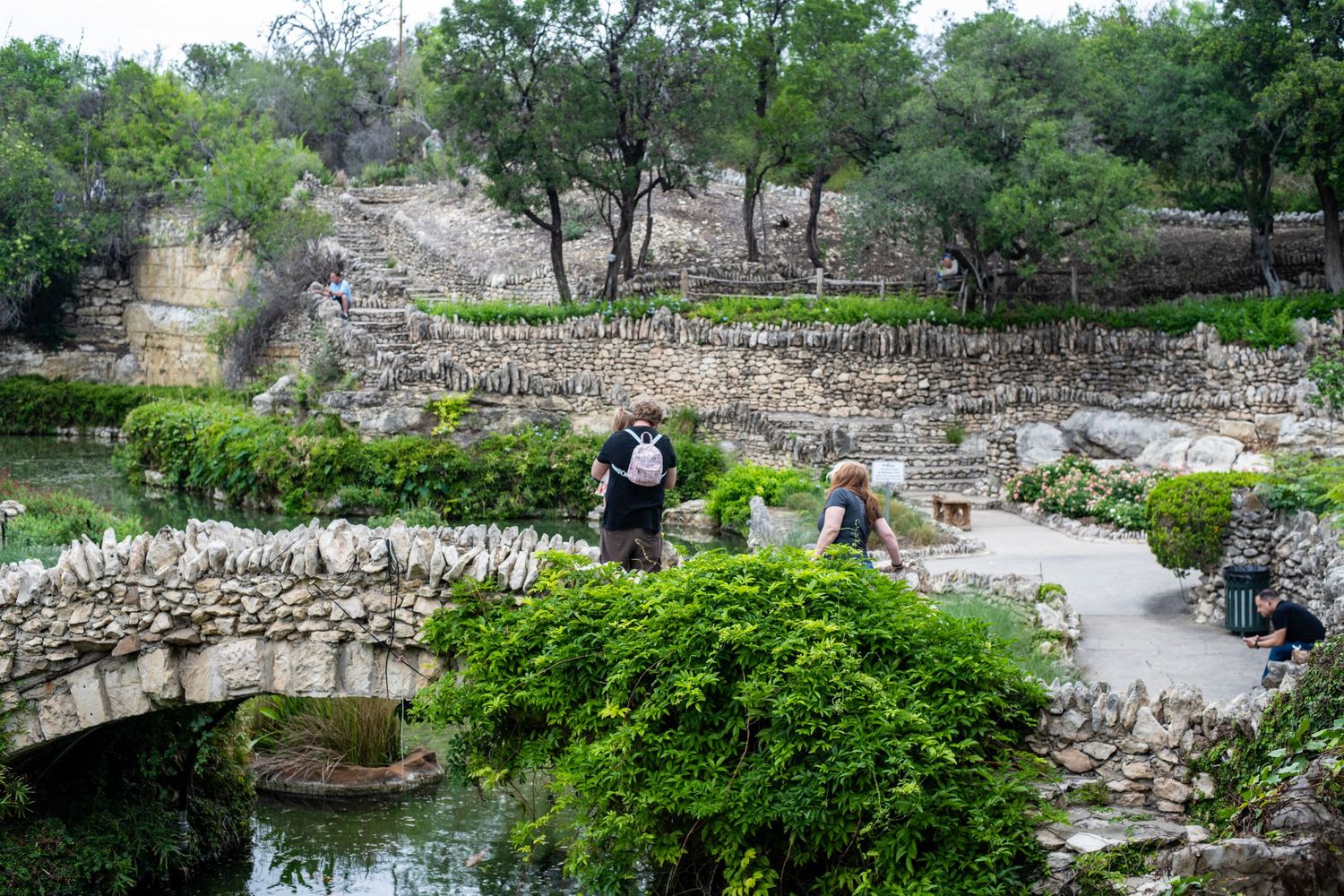Visitors explore a lush stone garden with a pond, rock walls, and greenery on a cloudy day.