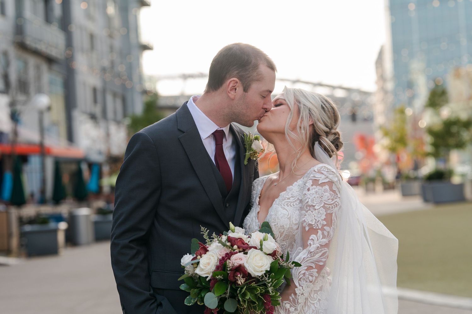 Bride and groom share a romantic kiss outdoors, bride holding red and white floral bouquet in elegant lace wedding dress.