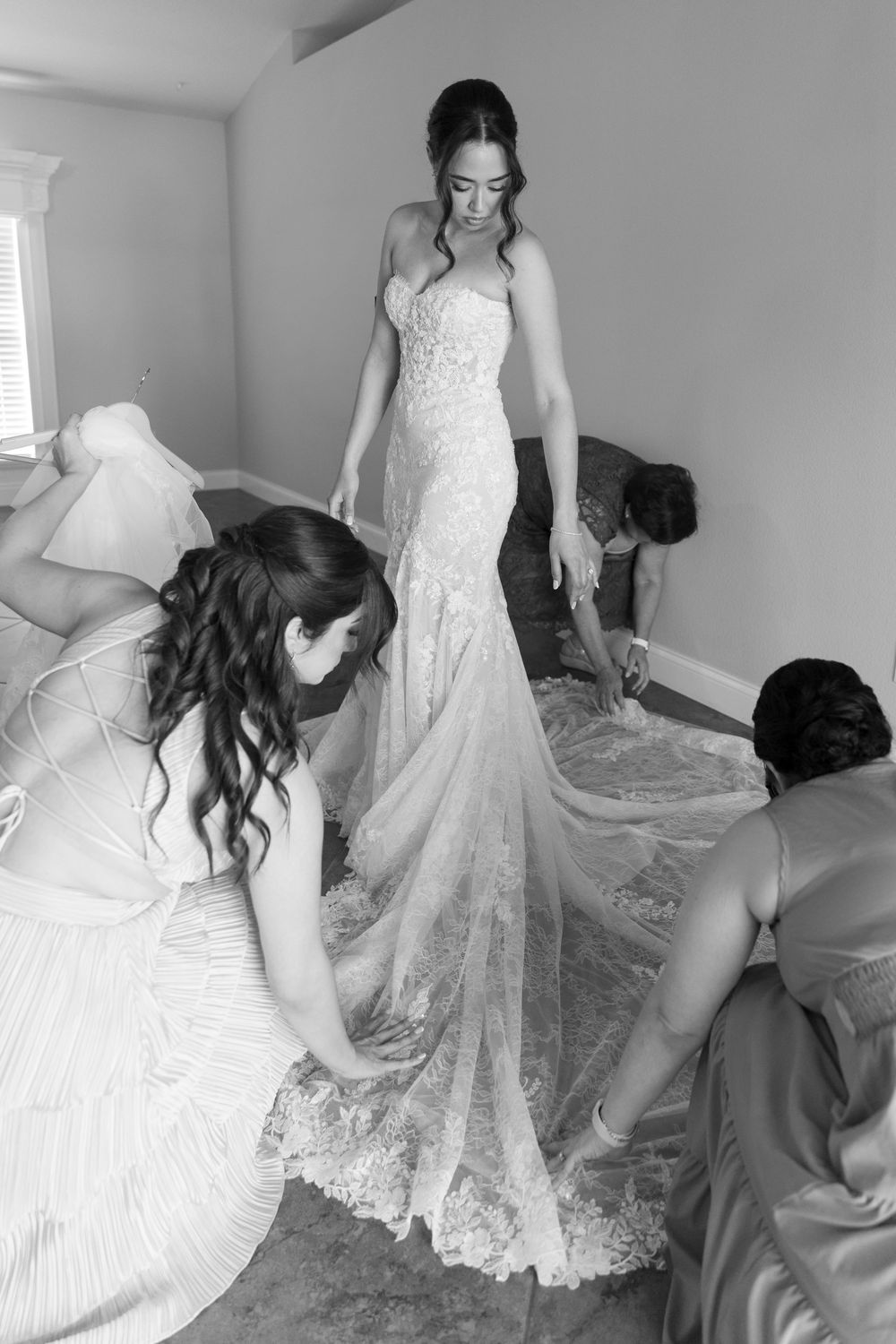 Bride in lace wedding gown being helped by bridesmaids arranging her train in a black and white photo.