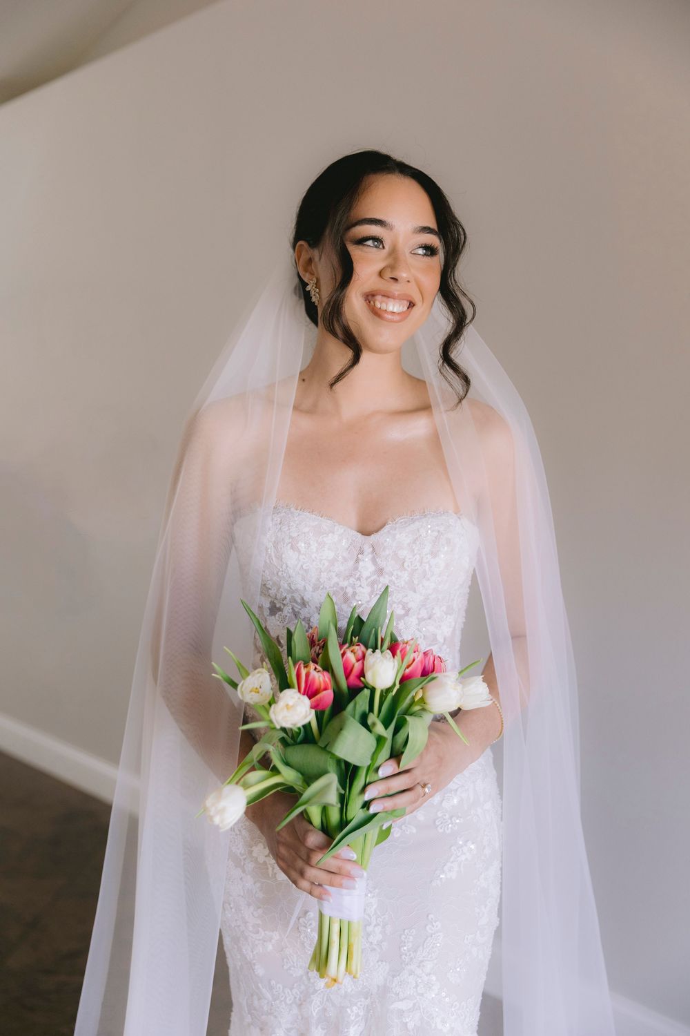 Smiling bride in strapless lace gown and veil holds colorful tulip bouquet indoors.