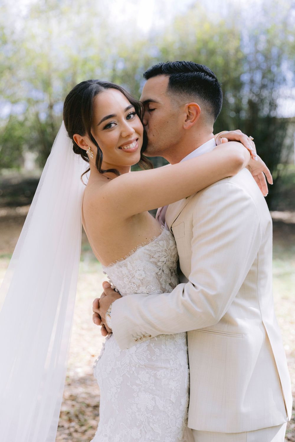 Bride in lace gown and veil smiles as groom in ivory suit kisses her cheek outdoors.