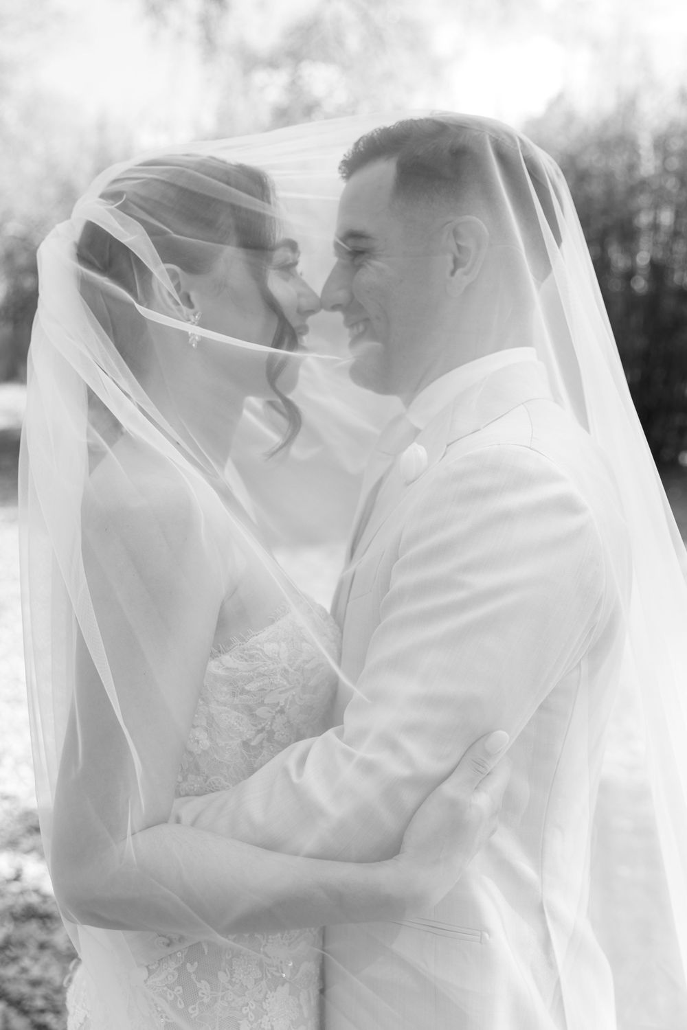 Black and white photo of bride and groom sharing intimate moment under wedding veil, smiling closely together outdoors.