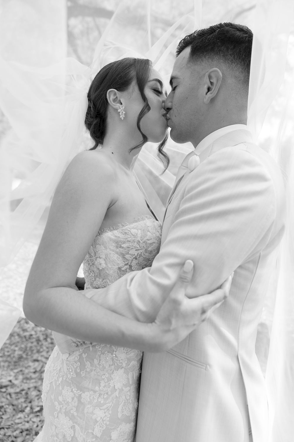 Black and white photo of a bride and groom sharing a romantic kiss under a flowing veil on their wedding day.