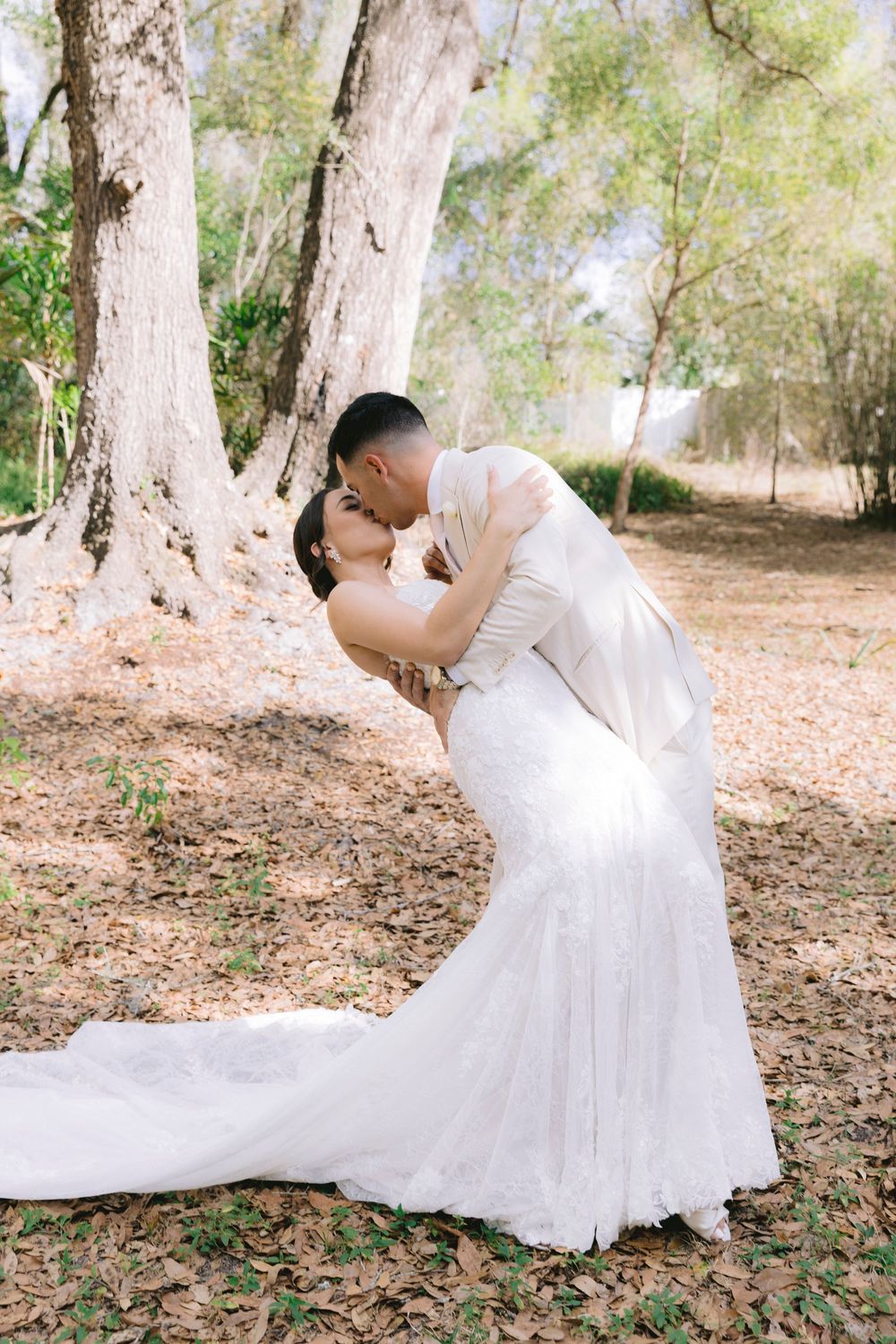 Groom dips bride in white lace mermaid gown for a romantic kiss among trees with autumn leaves on ground.