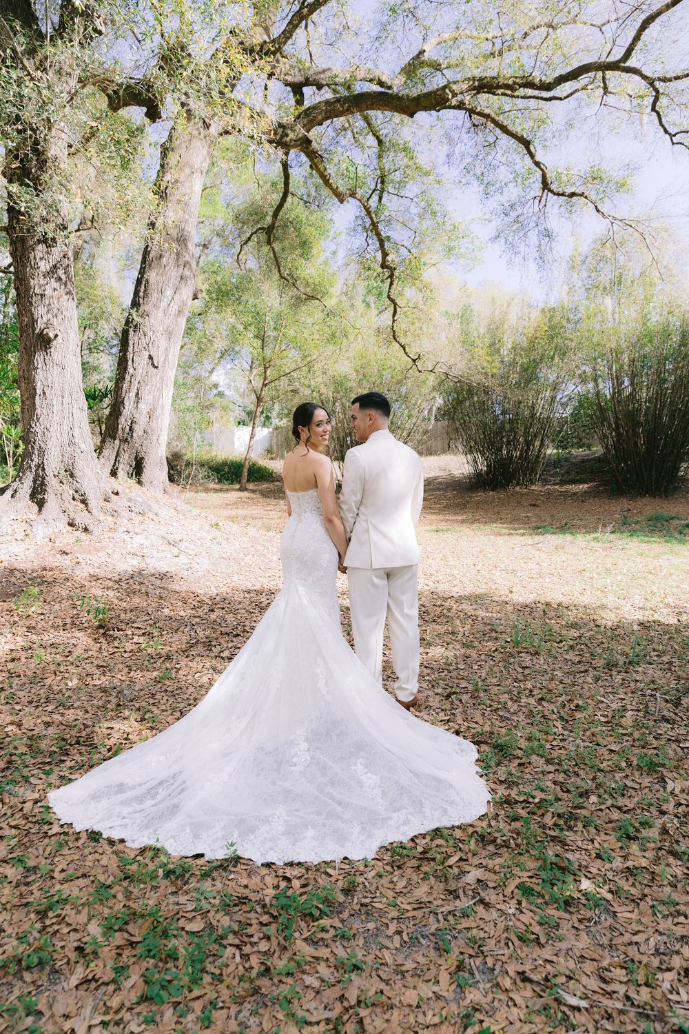 Bride in lace mermaid gown with long train stands with groom in beige suit under large trees in outdoor setting.