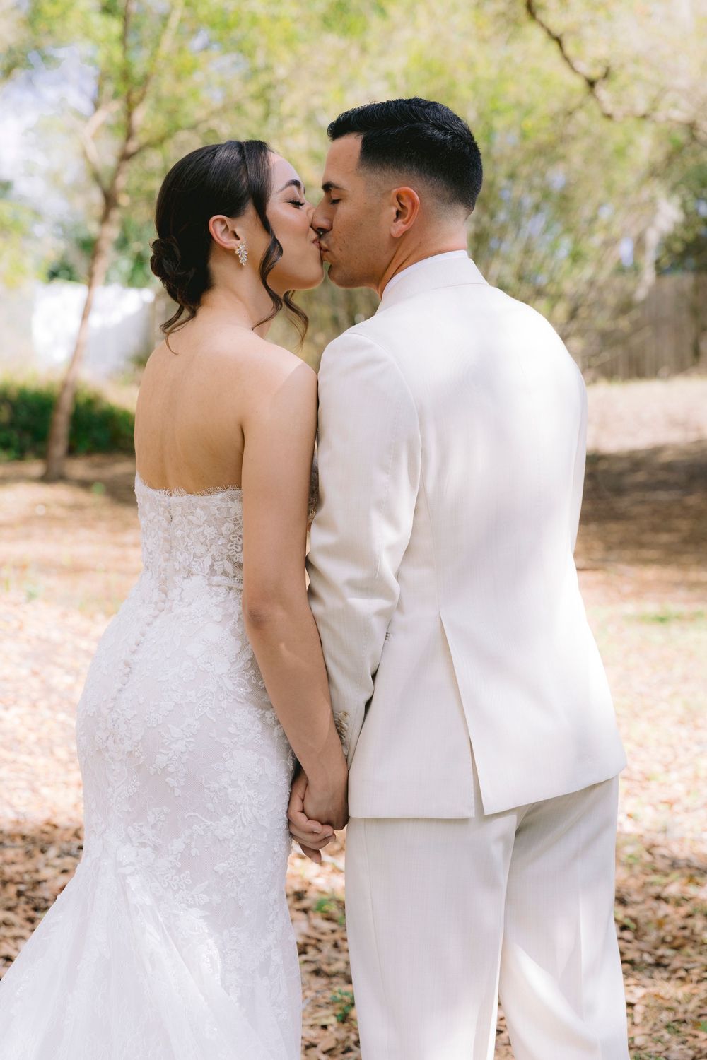 Bride and groom kiss while holding hands outdoors, wearing white wedding attire.