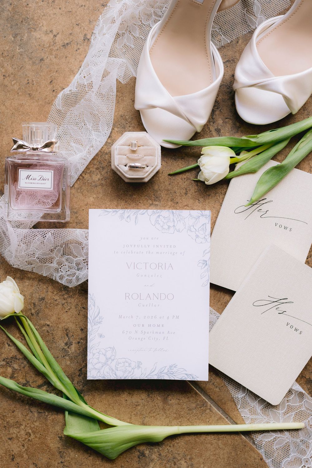 Elegant wedding flat lay with white heels, Miss Dior perfume, ring box, tulips, and Victoria and Rolando invitation suite.