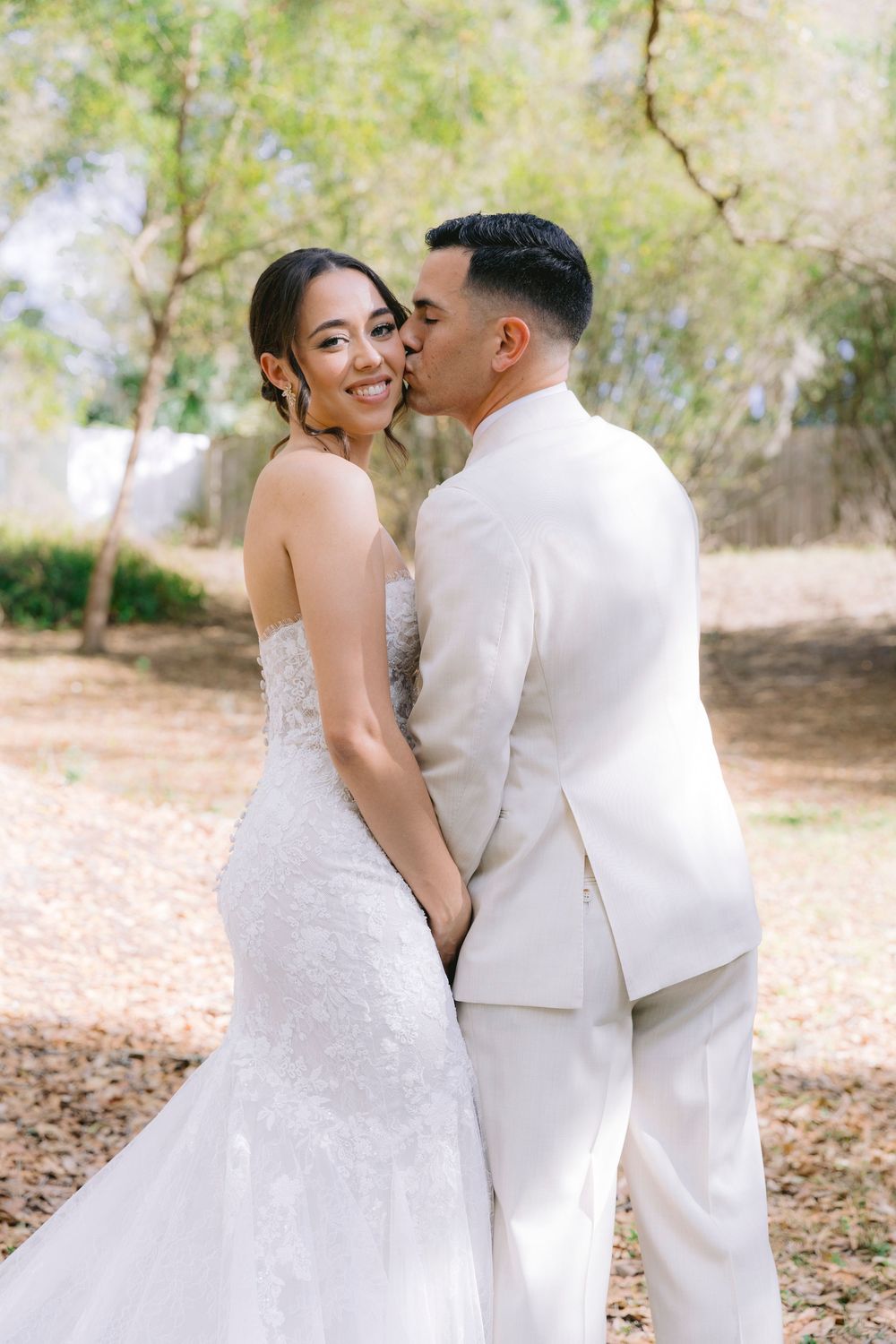 Groom in white suit kisses bride in strapless lace wedding dress while holding hands outdoors.
