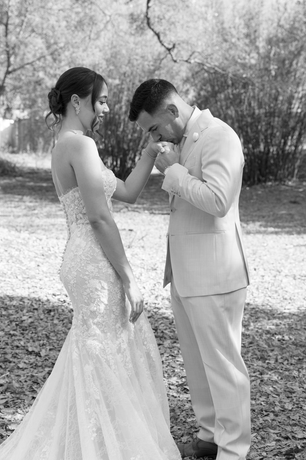 Black and white photo of a groom kissing his bride's hand outdoors on their wedding day.