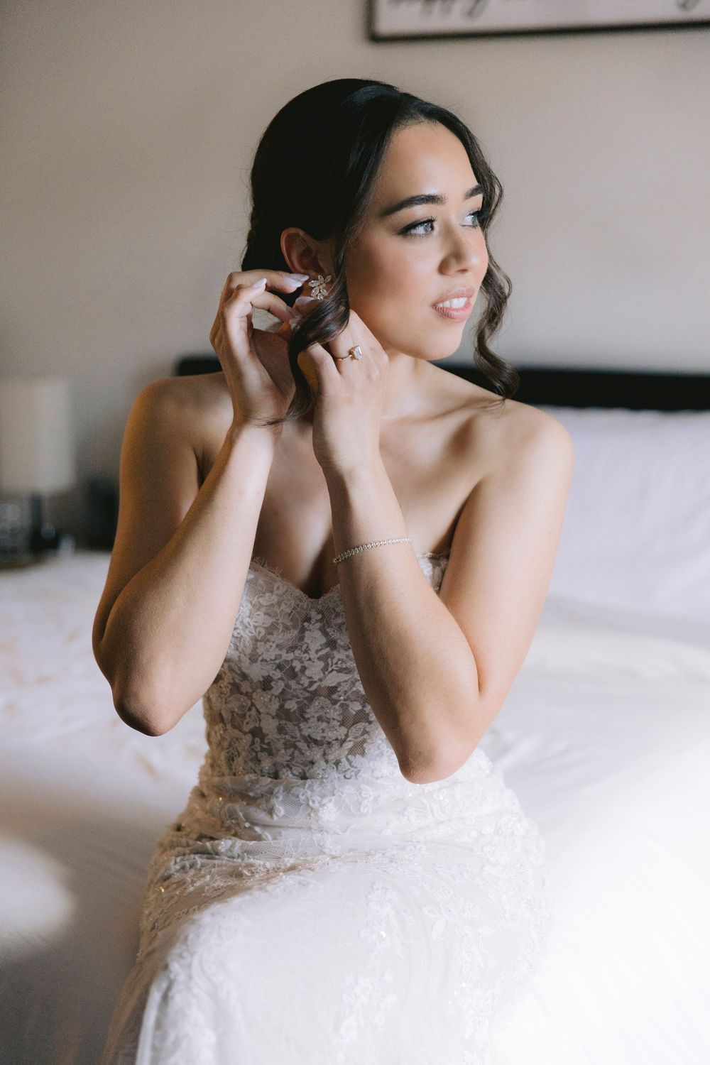 Bride in lace wedding gown sitting on bed putting on earrings while getting ready on her wedding day.