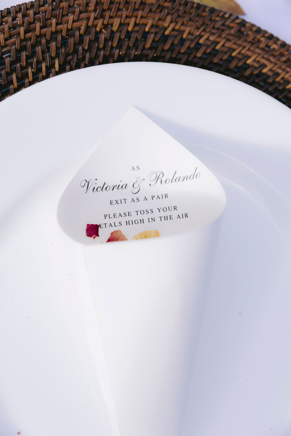 Wedding petal toss cone on white plate with Victoria & Rolando exit instructions, placed on woven charger.