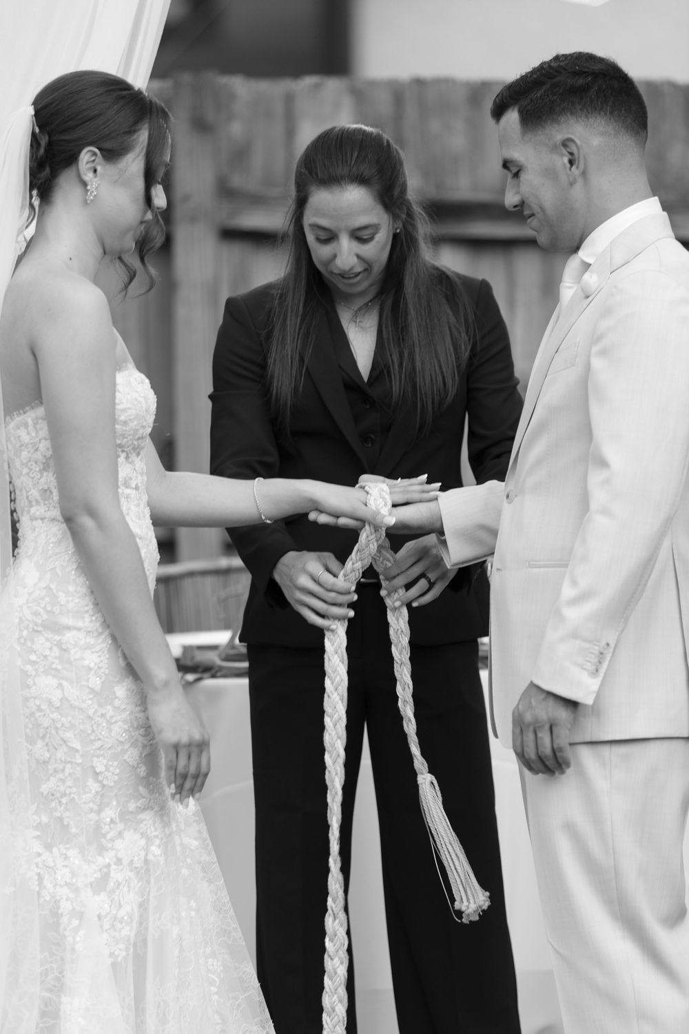 Black and white photo of a wedding officiant performing a hand-fasting ceremony with a bride in lace gown and groom in white suit.