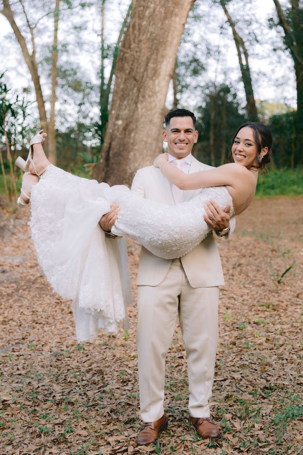 Groom in beige suit carries smiling bride in white lace wedding dress outdoors among autumn leaves and trees.