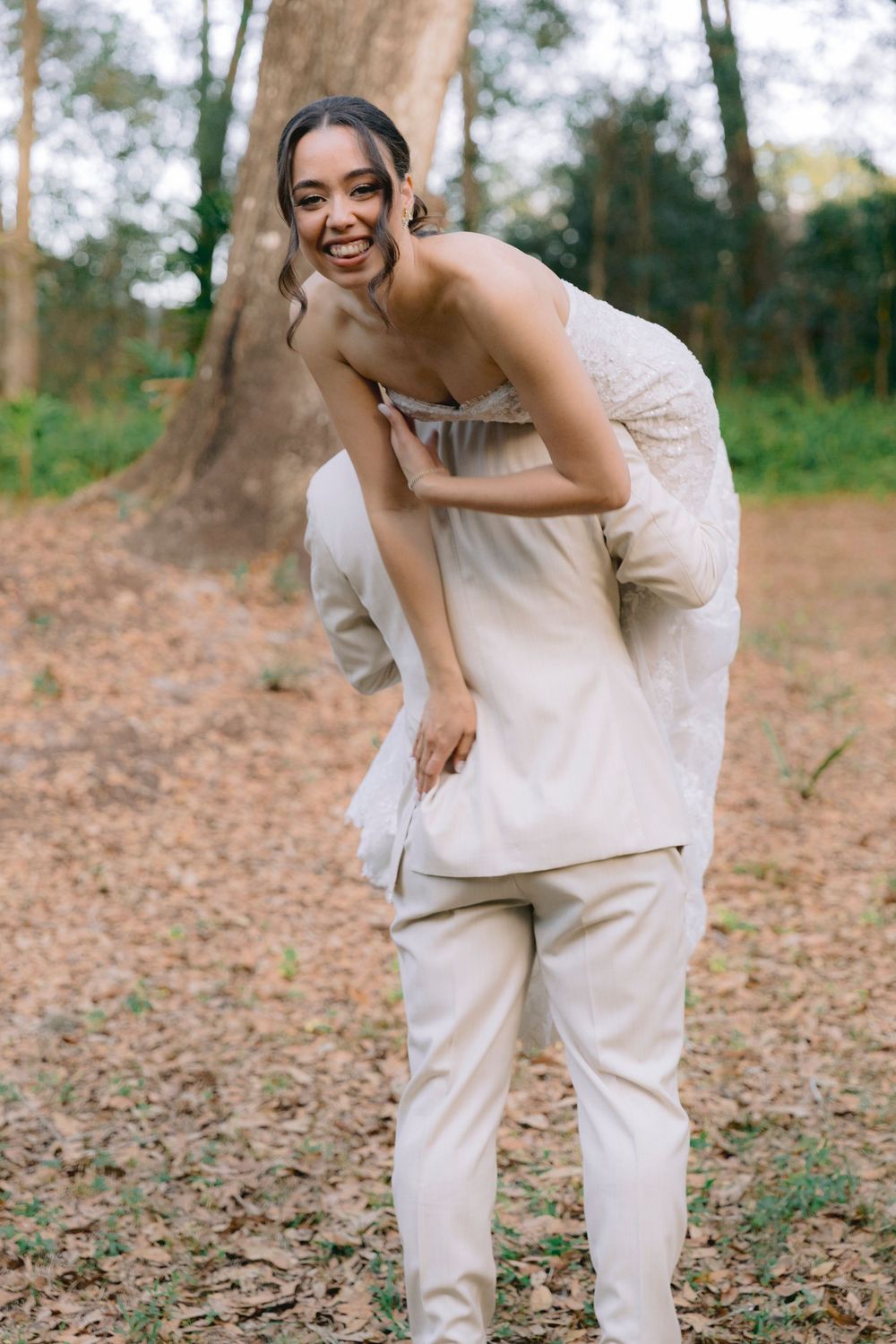 Laughing bride in strapless white gown draped over groom's shoulder in playful wedding portrait outdoors.