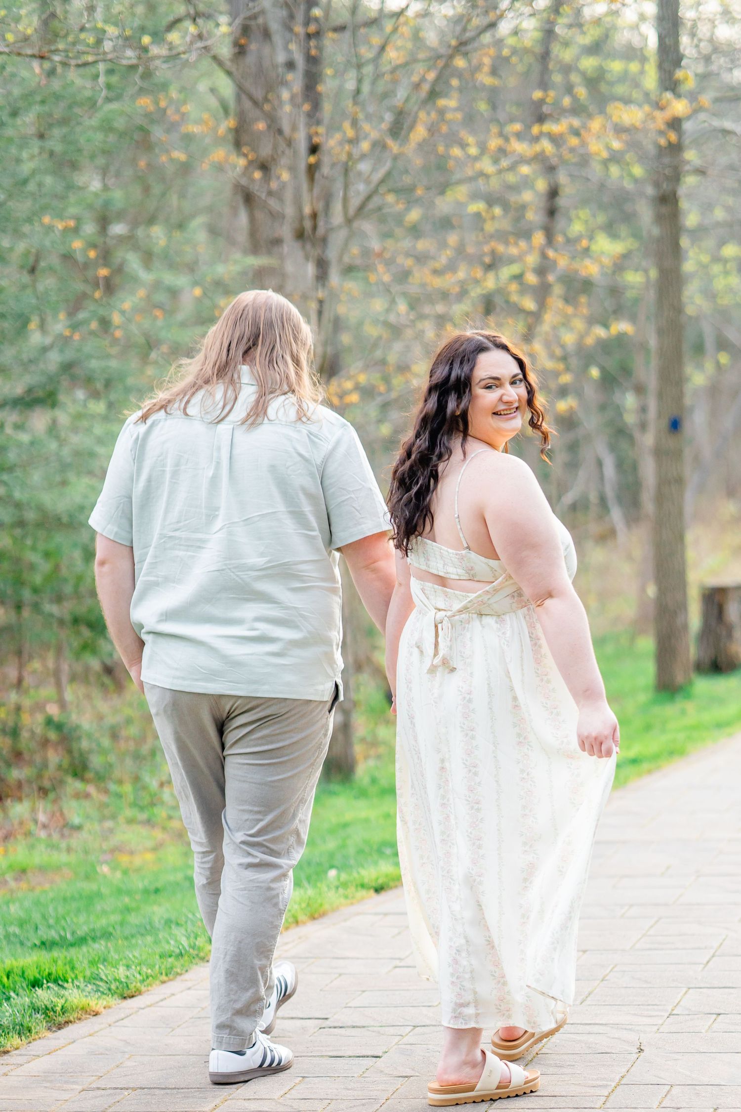 Couple walking on a brick path, woman in white dress glancing back smiling, man in mint shirt, surrounded by autumn trees.
