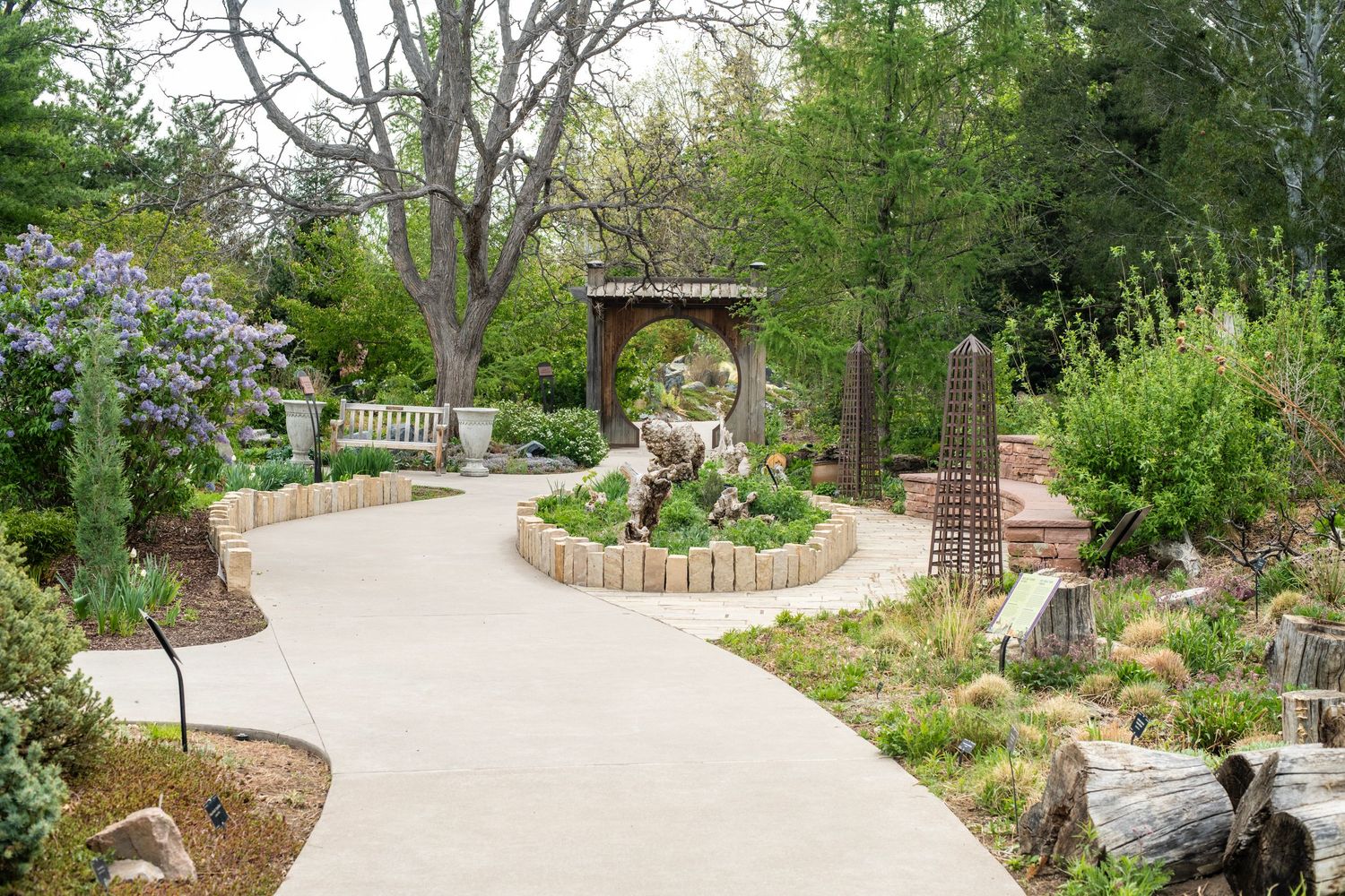 Winding concrete path through a lush botanical garden with decorative arch, sculptures, and colorful plantings.