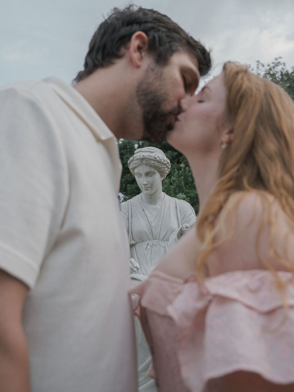 A couple shares a romantic kiss in front of a classical stone statue in a lush garden setting.
