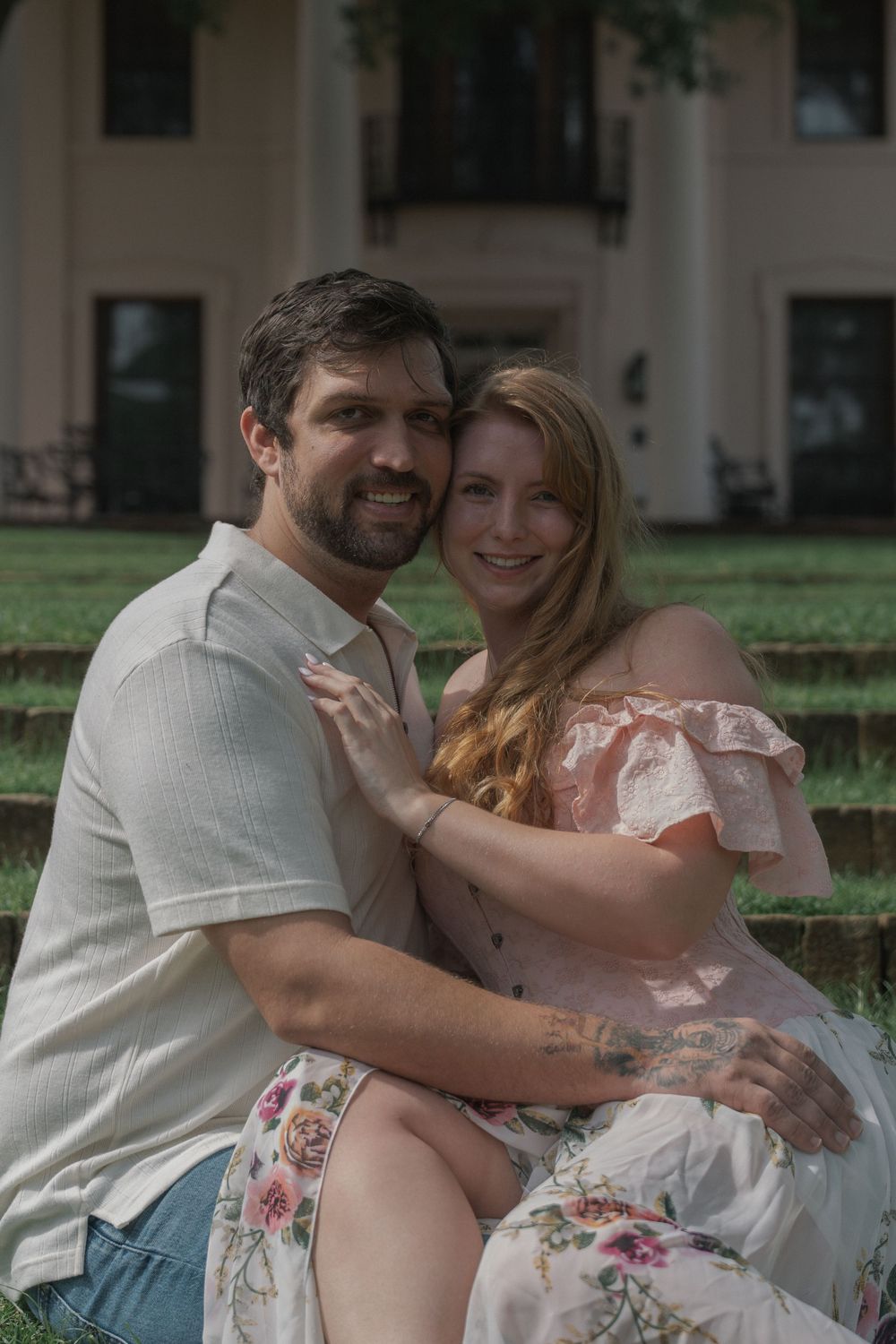 A smiling couple in casual elegant attire sits closely together on steps outside a historic mansion.
