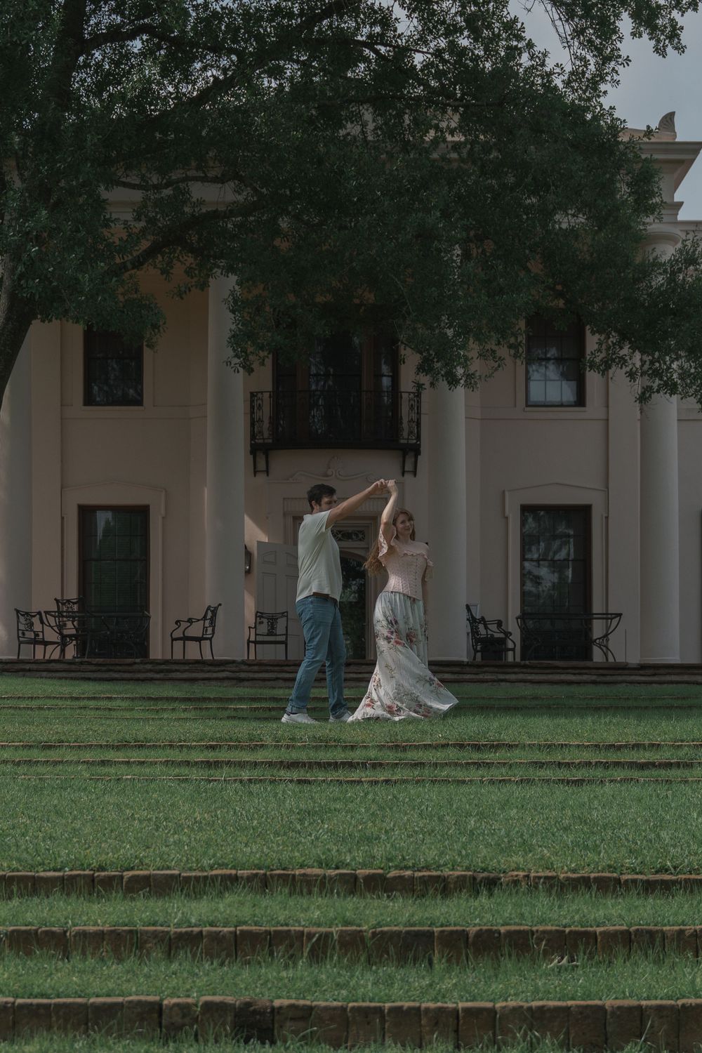 A couple dances joyfully on a manicured lawn in front of a grand white colonial-style estate.