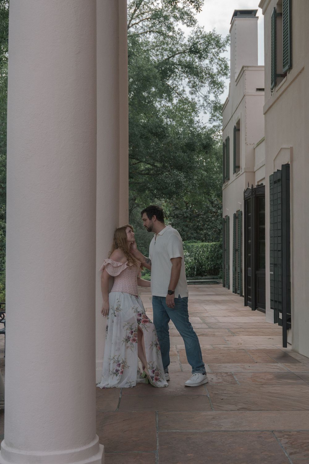 A couple shares an intimate moment between white columns on the covered walkway of an elegant estate.